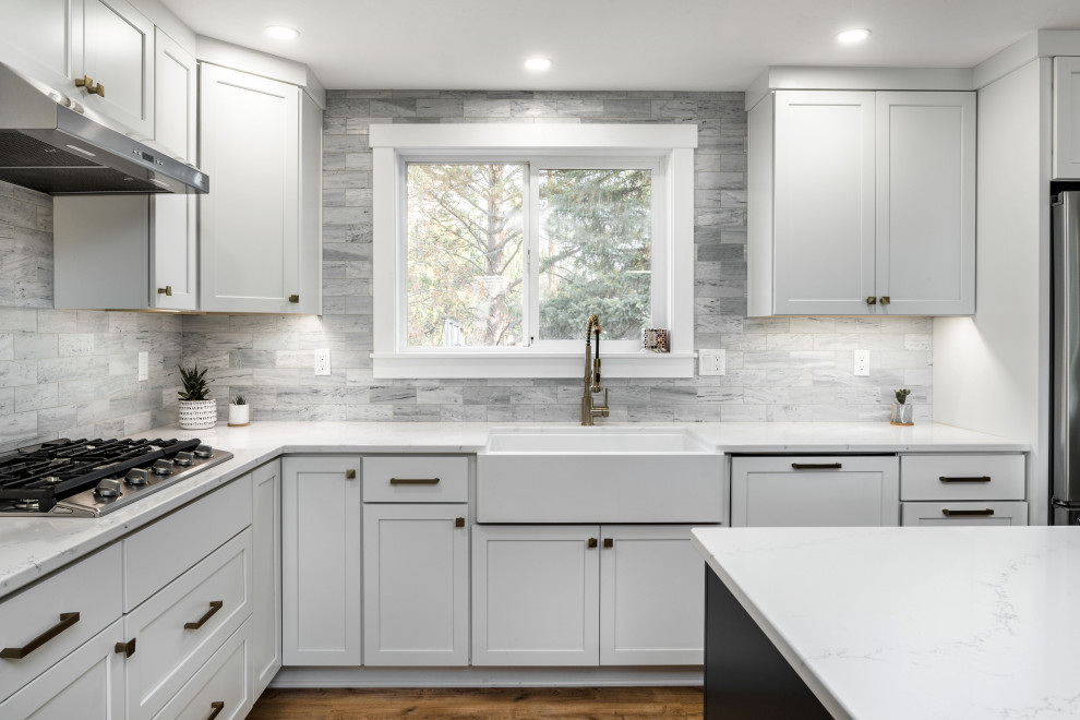 Design ideas for a medium sized contemporary l-shaped open plan kitchen in Boise with a belfast sink, shaker cabinets, white cabinets, engineered stone countertops, grey splashback, marble splashback, stainless steel appliances, laminate floors, an island, brown floors and white worktops.