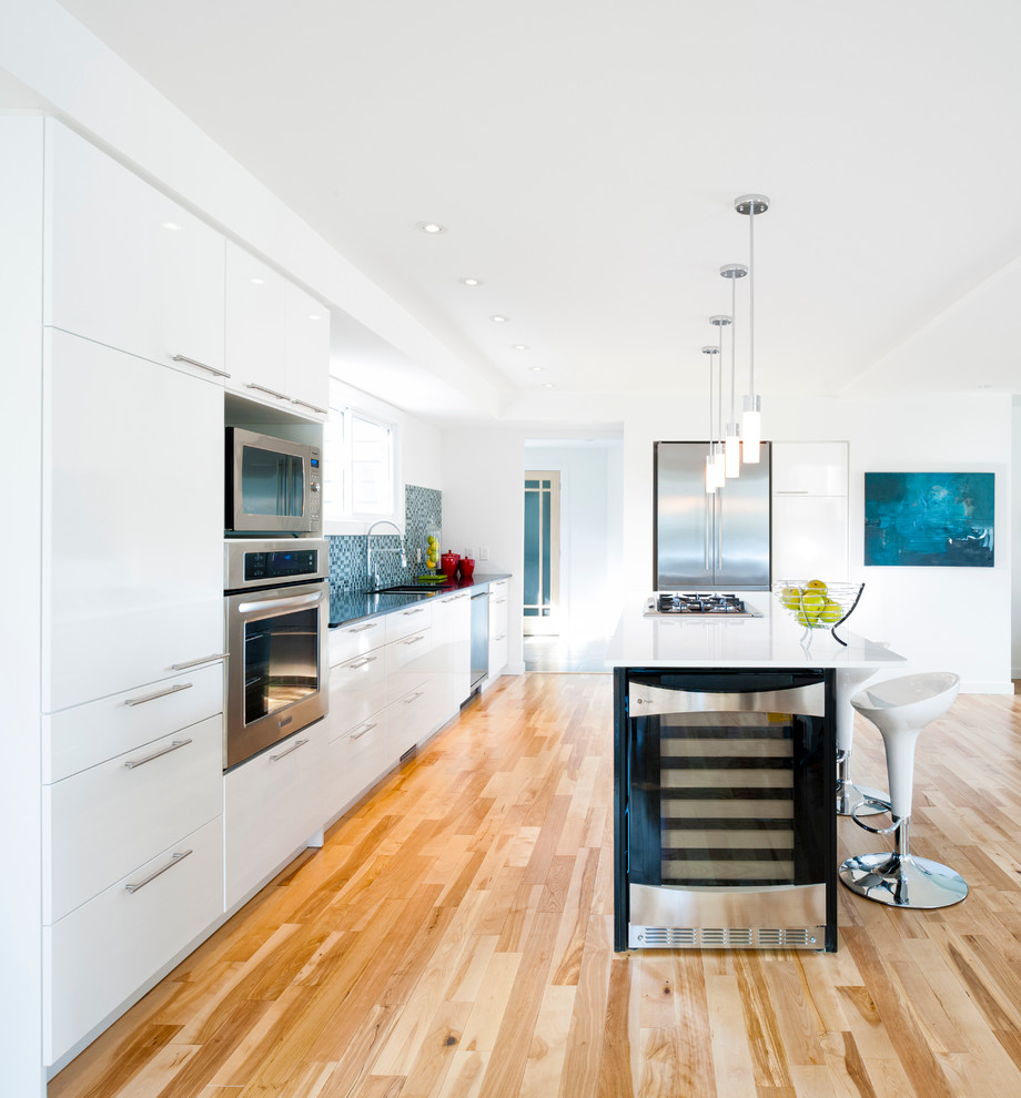Large contemporary galley open plan kitchen in Ottawa with flat-panel cabinets, white cabinets, stainless steel appliances, light hardwood floors, with island, an undermount sink, quartz benchtops, multi-coloured splashback and glass tile splashback.