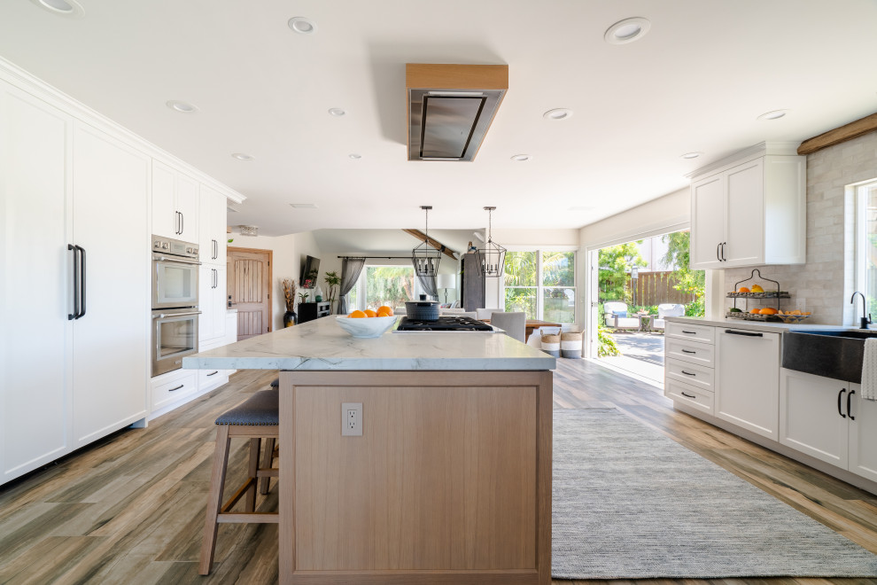 Inspiration for an expansive country galley open plan kitchen in San Diego with a farmhouse sink, recessed-panel cabinets, light wood cabinets, quartzite benchtops, beige splashback, ceramic splashback, stainless steel appliances, porcelain floors, with island, brown floor, white benchtop and exposed beam.