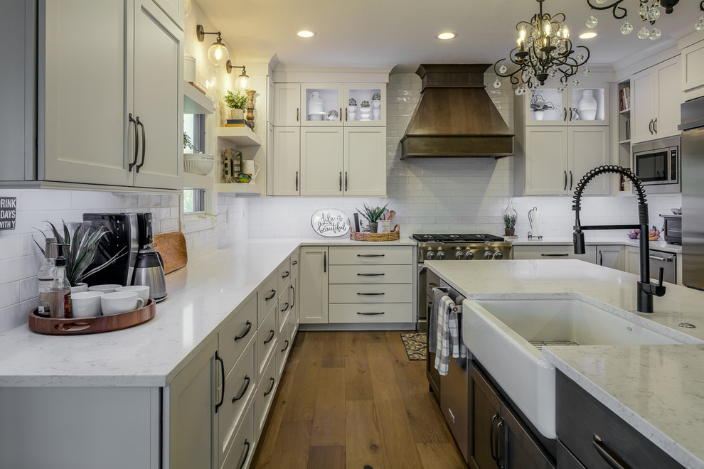 U-shaped brown floor eat-in kitchen photo with a farmhouse sink, shaker cabinets, white cabinets, quartz countertops, white backsplash, subway tile backsplash, stainless steel appliances, an island and white countertops