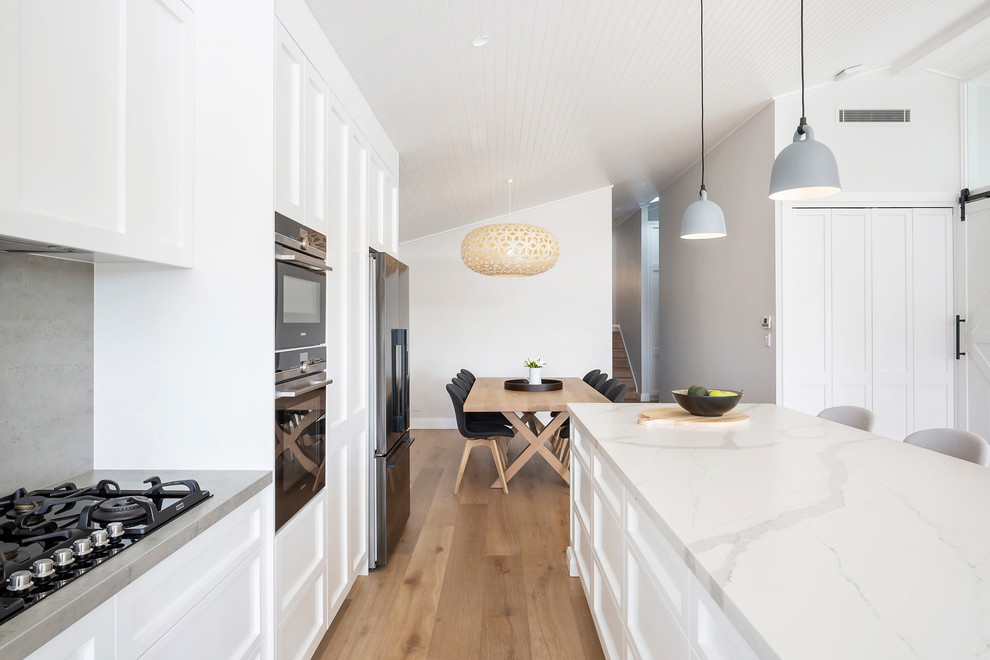 Photo of a mid-sized beach style l-shaped open plan kitchen in Sydney with a double-bowl sink, shaker cabinets, white cabinets, quartz benchtops, grey splashback, stainless steel appliances, medium hardwood floors, with island, brown floor and white benchtop.