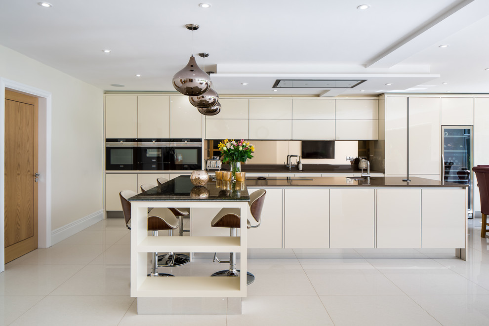This is an example of an expansive contemporary u-shaped open plan kitchen in Hertfordshire with a drop-in sink, flat-panel cabinets, white cabinets, glass benchtops, metallic splashback, mirror splashback, stainless steel appliances, slate floors, multiple islands, yellow floor and brown benchtop.