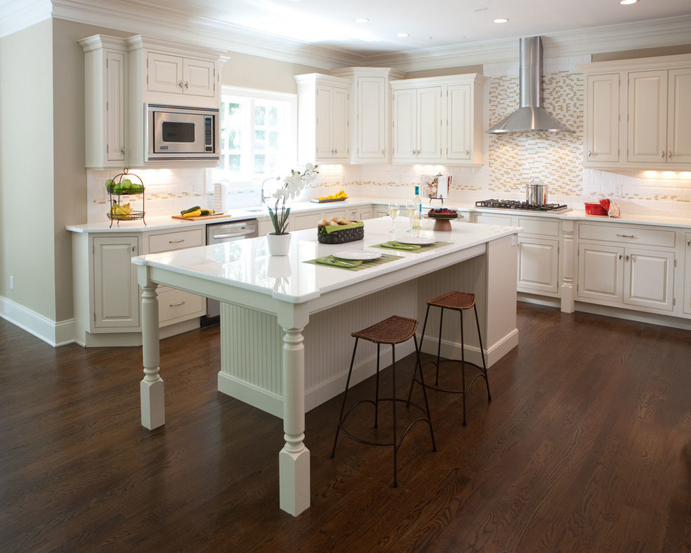 Large transitional l-shaped dark wood floor and brown floor eat-in kitchen photo with a drop-in sink, raised-panel cabinets, white cabinets, solid surface countertops, white backsplash, ceramic backsplash, stainless steel appliances and an island