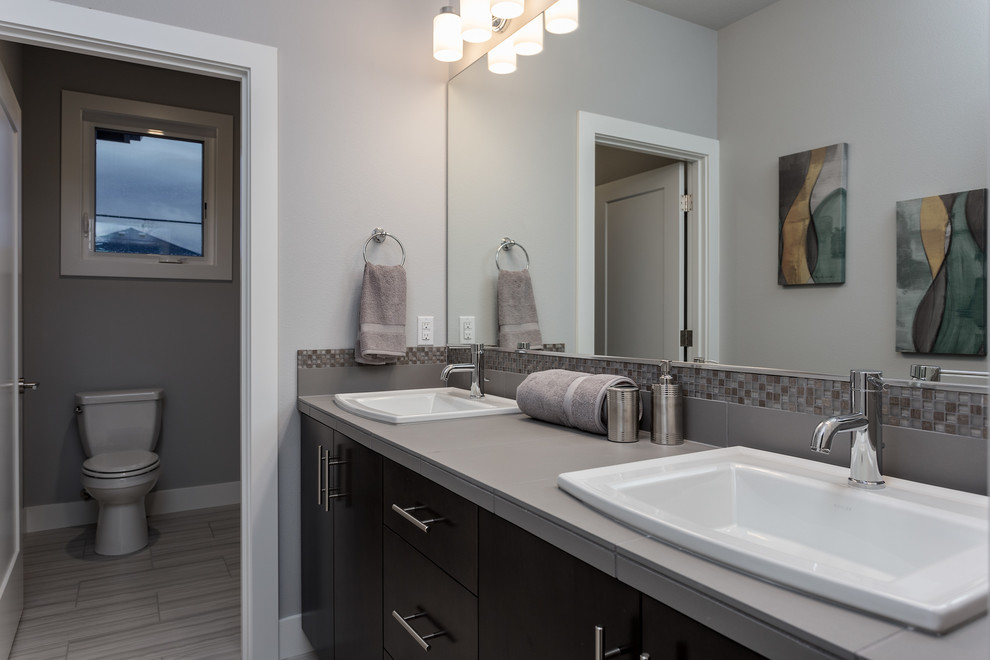 Inspiration for a large transitional master bathroom in Portland with flat-panel cabinets, dark wood cabinets, a drop-in tub, a corner shower, a two-piece toilet, gray tile, glass sheet wall, grey walls, ceramic floors, a drop-in sink, tile benchtops, grey floor and a hinged shower door.