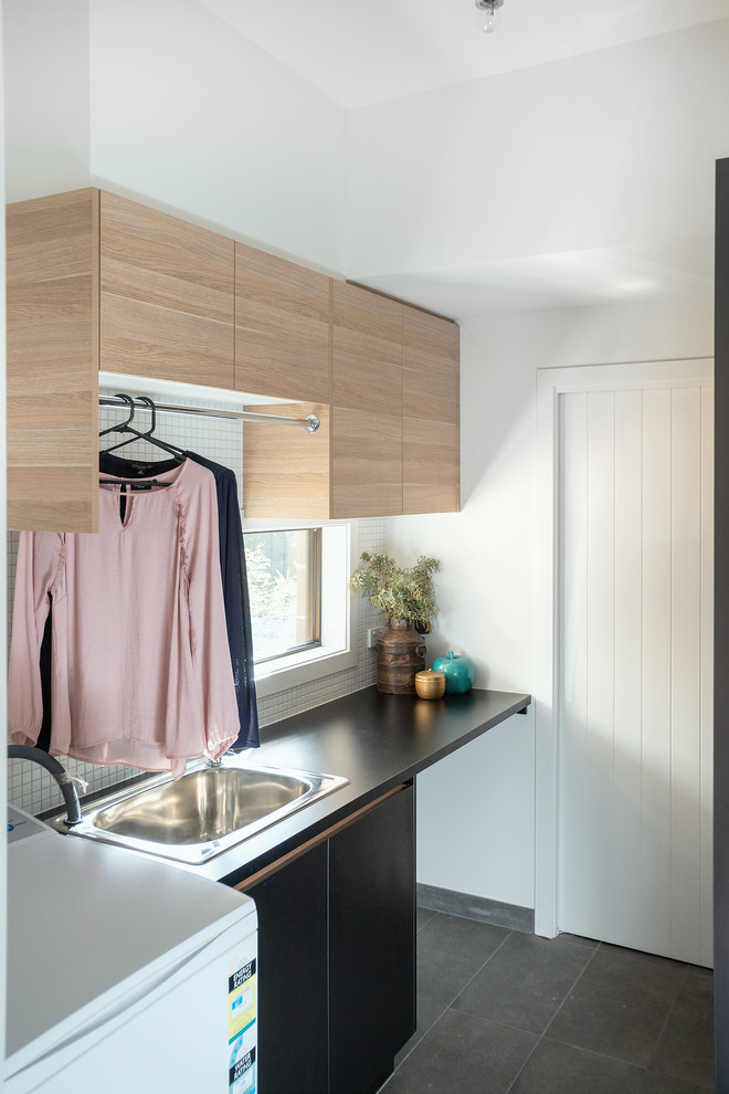 This is an example of a small beach style galley dedicated laundry room in Other with a drop-in sink, black cabinets, laminate benchtops, white walls, porcelain floors, grey floor and black benchtop.