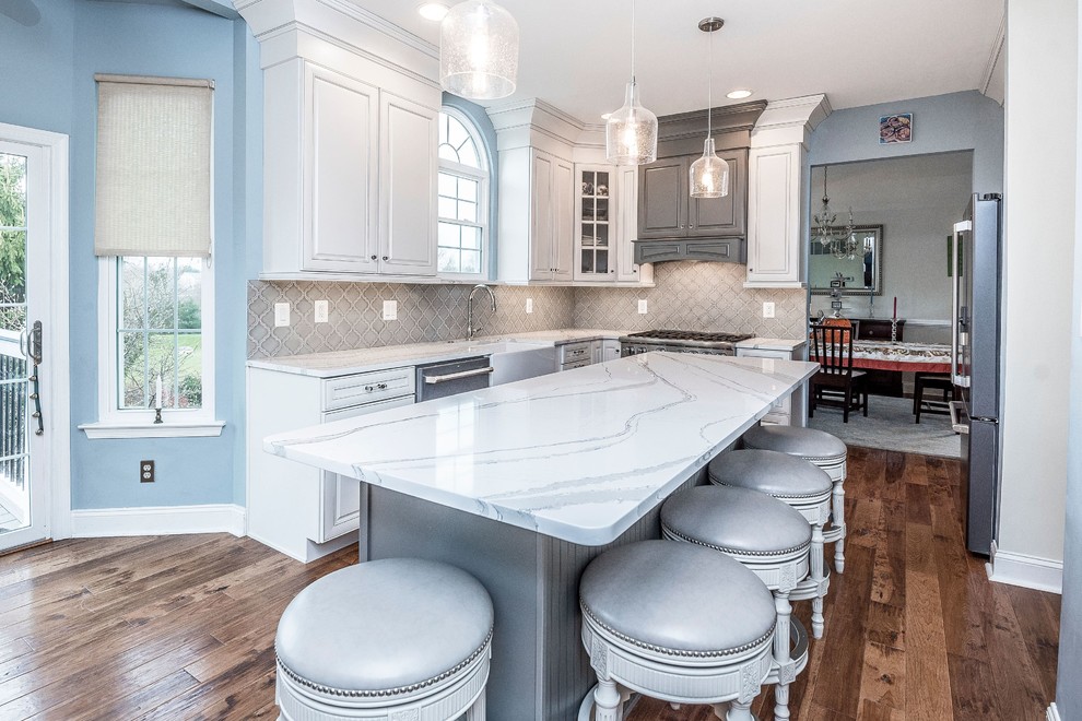 Large contemporary u-shaped open plan kitchen in Philadelphia with a farmhouse sink, raised-panel cabinets, white cabinets, marble benchtops, grey splashback, ceramic splashback, stainless steel appliances, medium hardwood floors, with island, brown floor and white benchtop.