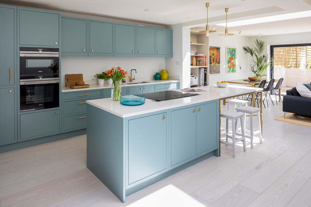 Mid-sized contemporary single-wall open plan kitchen in London with a double-bowl sink, flat-panel cabinets, turquoise cabinets, quartzite benchtops, stainless steel appliances, light hardwood floors, with island, grey floor and white benchtop.