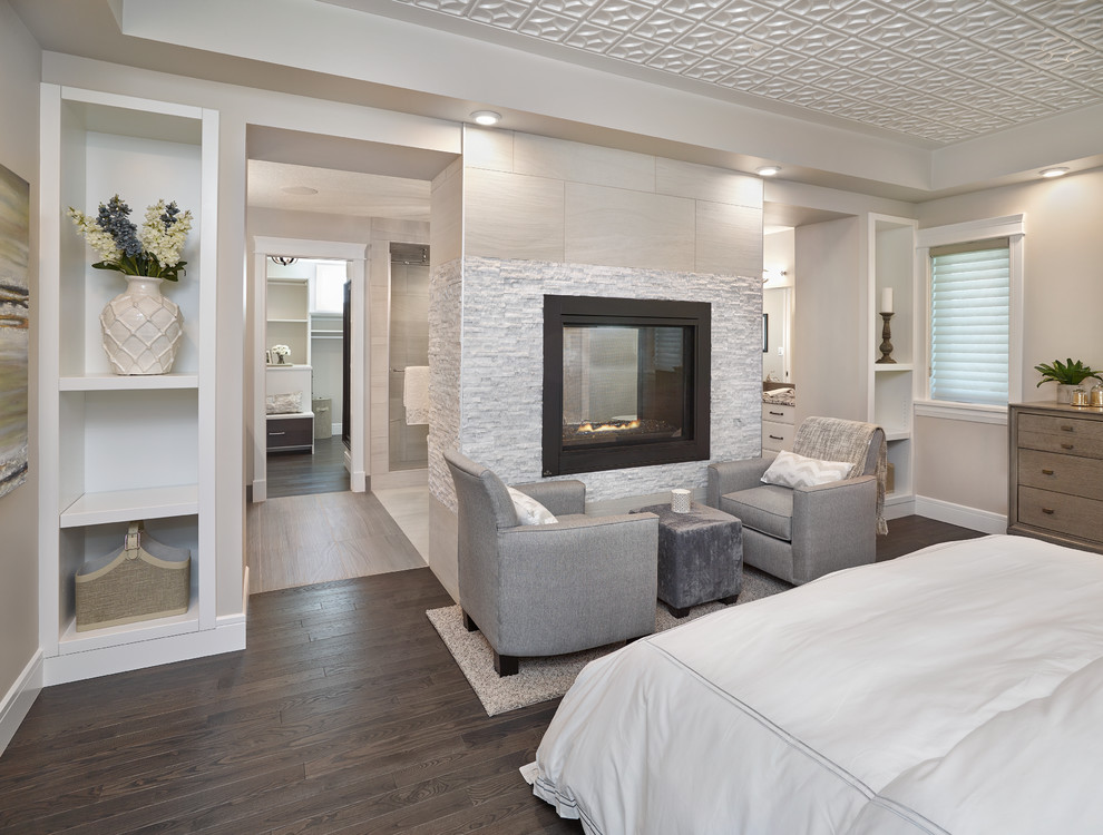 This is an example of a transitional master bedroom in Edmonton with beige walls, dark hardwood floors, a two-sided fireplace and a tile fireplace surround.