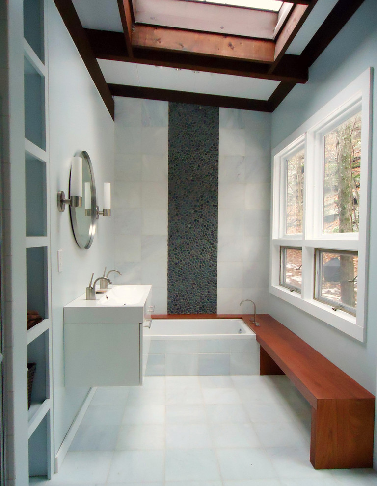 Midcentury bathroom in New York with a wall-mount sink, flat-panel cabinets, white cabinets, solid surface benchtops, a drop-in tub, an alcove shower, gray tile, pebble tile, grey walls and marble floors.