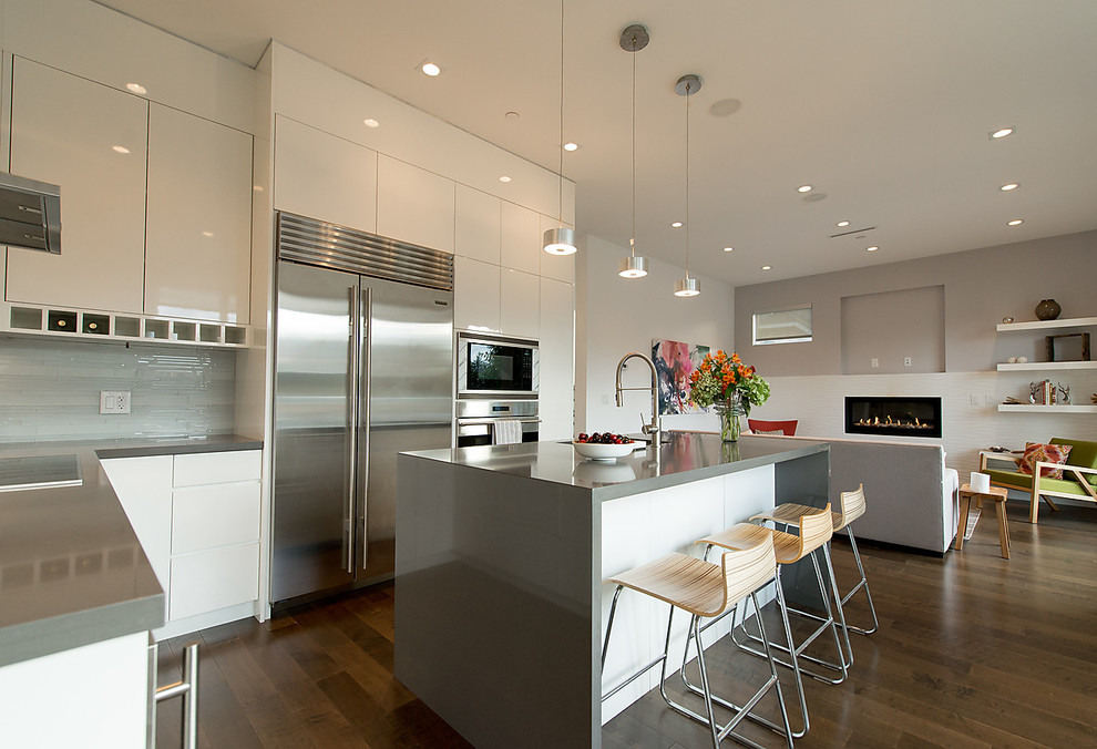 Photo of a mid-sized modern l-shaped open plan kitchen in Vancouver with an undermount sink, flat-panel cabinets, white cabinets, solid surface benchtops, grey splashback, stainless steel appliances, medium hardwood floors, with island, brown floor and grey benchtop.