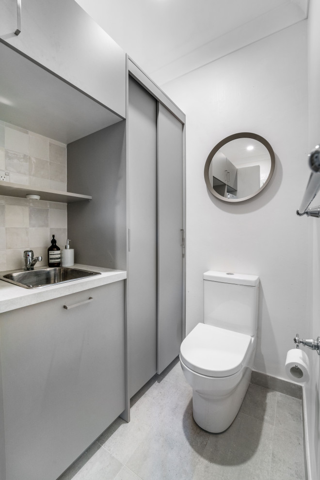 This is an example of a mid-sized transitional single-wall powder room in Sydney with a drop-in sink, flat-panel cabinets, grey cabinets, quartz benchtops, multi-coloured splashback, ceramic splashback, multi-coloured walls, ceramic floors, an integrated washer and dryer, grey floor and white benchtop.