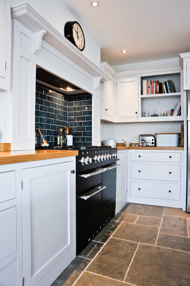 This is an example of a large traditional u-shaped eat-in kitchen in Gloucestershire with a double-bowl sink, shaker cabinets, granite benchtops, black splashback, stone tile splashback, black appliances, travertine floors, with island, beige floor and black benchtop.