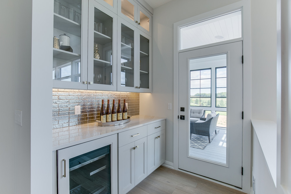Large transitional single-wall home bar in Minneapolis with an undermount sink, shaker cabinets, white cabinets, quartzite benchtops, multi-coloured splashback, light hardwood floors, beige floor and grey benchtop.
