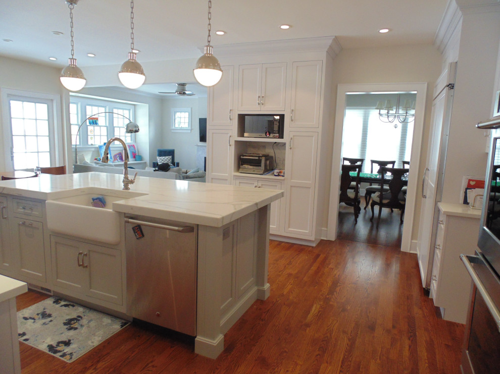 Example of a large transitional u-shaped medium tone wood floor and brown floor eat-in kitchen design with a farmhouse sink, recessed-panel cabinets, white cabinets, quartz countertops, white backsplash, subway tile backsplash, stainless steel appliances, an island and white countertops