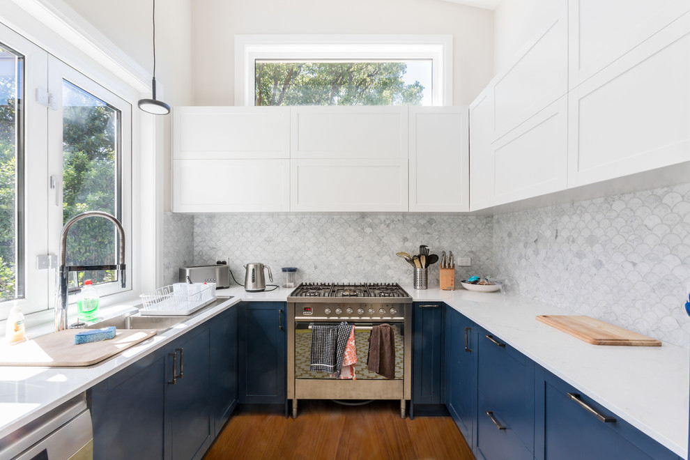 Design ideas for a transitional u-shaped kitchen in Sydney with a double-bowl sink, shaker cabinets, blue cabinets, white splashback, mosaic tile splashback, stainless steel appliances, medium hardwood floors, no island, brown floor and white benchtop.