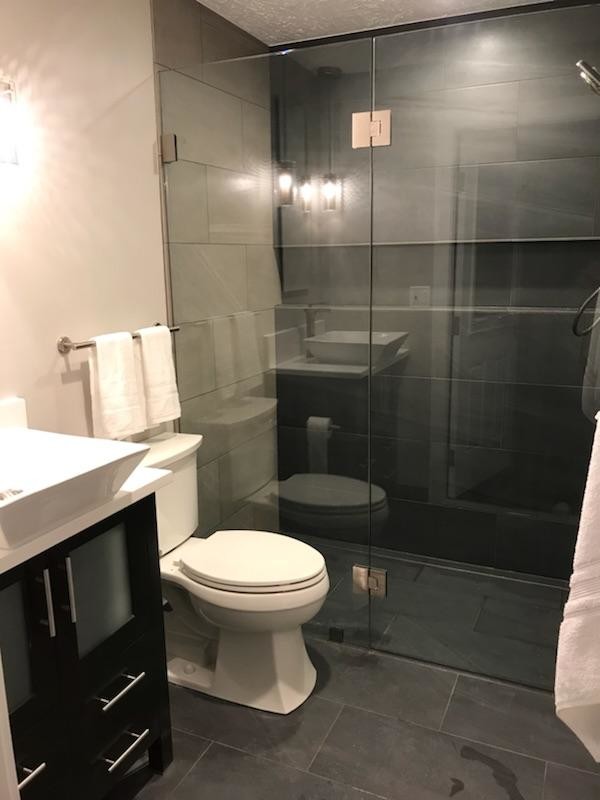 Photo of a small modern master bathroom in Portland with glass-front cabinets, dark wood cabinets, a curbless shower, a two-piece toilet, gray tile, ceramic tile, beige walls, ceramic floors, a vessel sink, engineered quartz benchtops, grey floor, a hinged shower door and white benchtops.