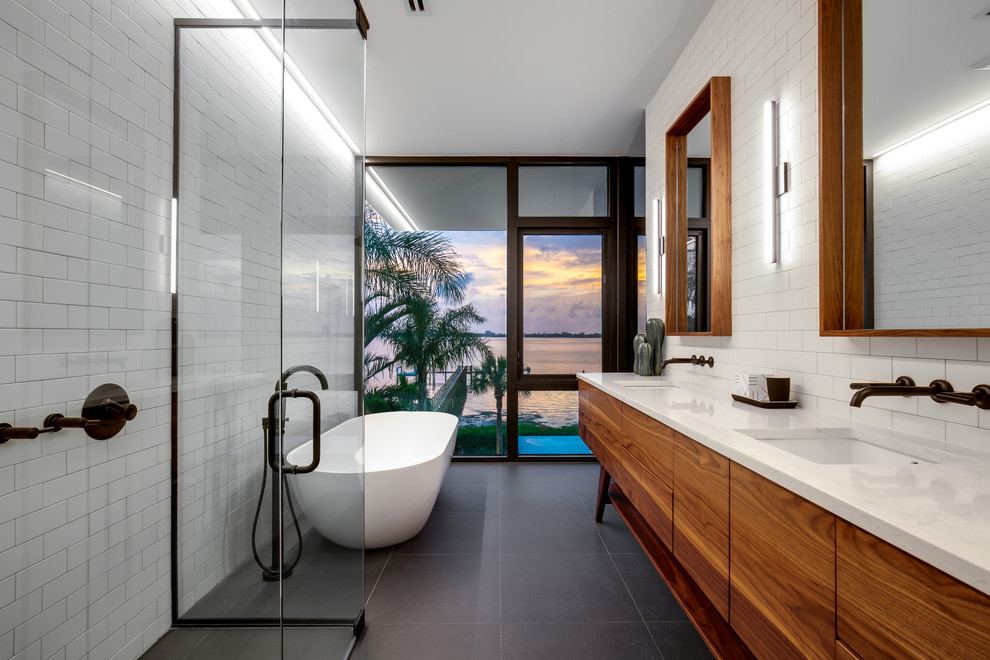 This is an example of a mid-sized modern master bathroom in Other with medium wood cabinets, a freestanding tub, a curbless shower, white tile, subway tile, porcelain floors, an undermount sink, engineered quartz benchtops, a hinged shower door, white benchtops, grey floor, white walls, flat-panel cabinets, a one-piece toilet, an enclosed toilet, a double vanity and a freestanding vanity.