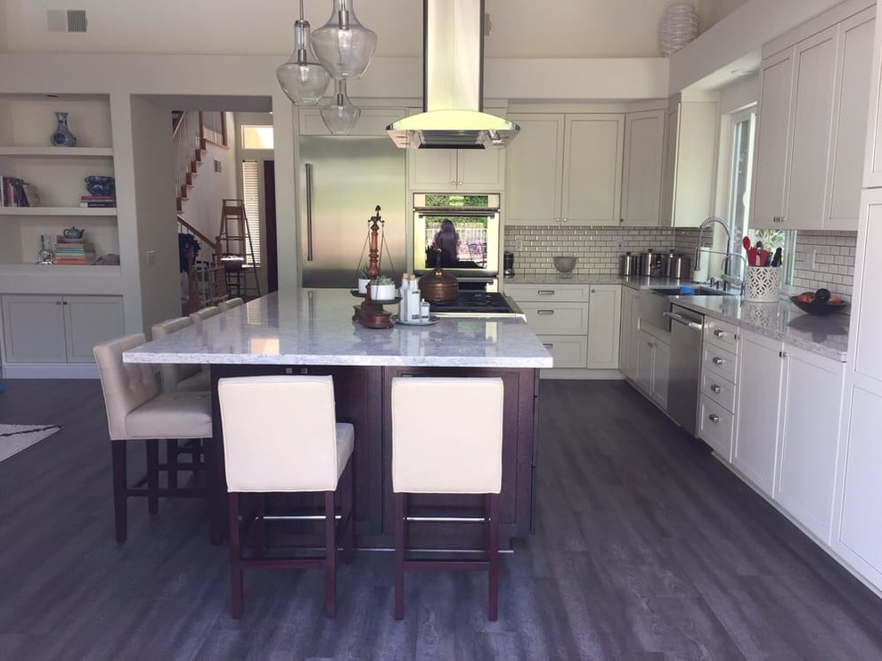 Photo of a large transitional l-shaped kitchen in San Diego with a farmhouse sink, shaker cabinets, white cabinets, granite benchtops, white splashback, subway tile splashback, stainless steel appliances, dark hardwood floors, with island and brown floor.
