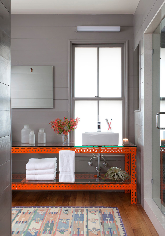На фото: ванная комната в стиле фьюжн с открытыми фасадами, оранжевыми фасадами, душем в нише, серыми стенами, паркетным полом среднего тона, настольной раковиной, стеклянной столешницей, коричневым полом, душем с распашными дверями, напольной тумбой и стенами из вагонки