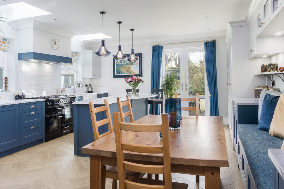 Large classic single-wall open plan kitchen in Cardiff with a belfast sink, shaker cabinets, blue cabinets, engineered stone countertops, white splashback, black appliances, light hardwood flooring, an island, brown floors and white worktops.