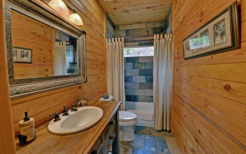 This is an example of a mid-sized country master bathroom in Other with furniture-like cabinets, distressed cabinets, an alcove tub, a shower/bathtub combo, a two-piece toilet, multi-coloured tile, slate, brown walls, slate floors, a drop-in sink, wood benchtops, multi-coloured floor and a shower curtain.