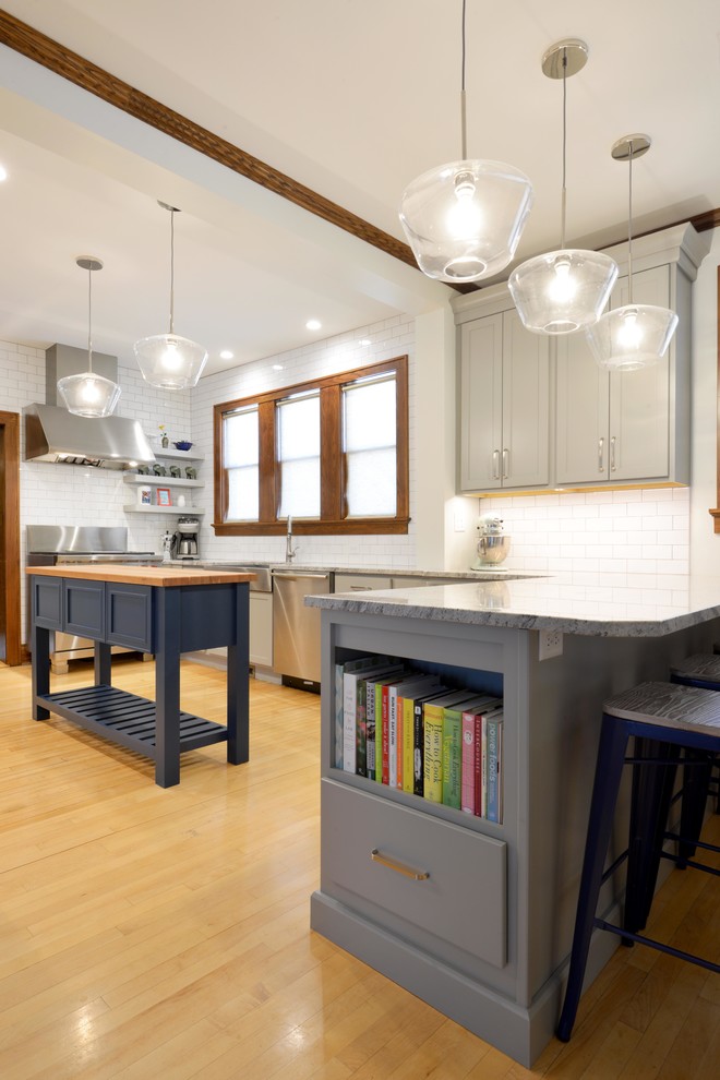 Example of a mid-sized transitional light wood floor and brown floor enclosed kitchen design with a farmhouse sink, shaker cabinets, gray cabinets, wood countertops, white backsplash, porcelain backsplash, stainless steel appliances, an island and brown countertops