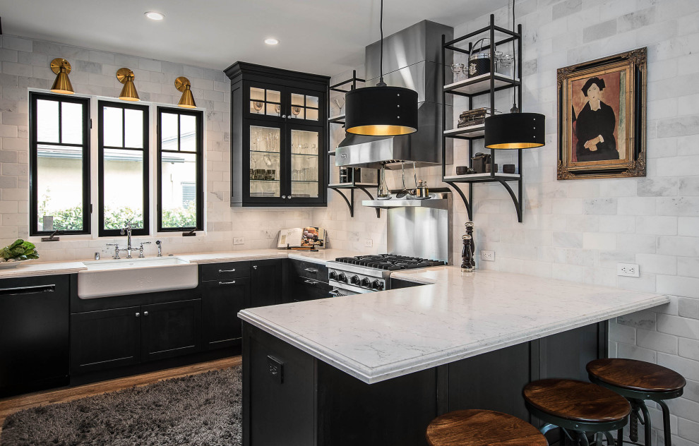 Photo of a mid-sized country u-shaped separate kitchen in Los Angeles with a farmhouse sink, a peninsula, grey benchtop, brown floor, glass-front cabinets, black cabinets, grey splashback, subway tile splashback, stainless steel appliances, light hardwood floors and quartz benchtops.