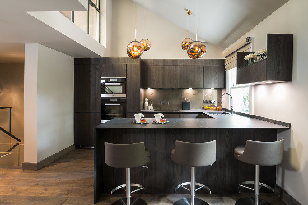 This is an example of a contemporary u-shaped kitchen in London with an undermount sink, flat-panel cabinets, dark wood cabinets, grey splashback, black appliances, dark hardwood floors, a peninsula, brown floor and black benchtop.