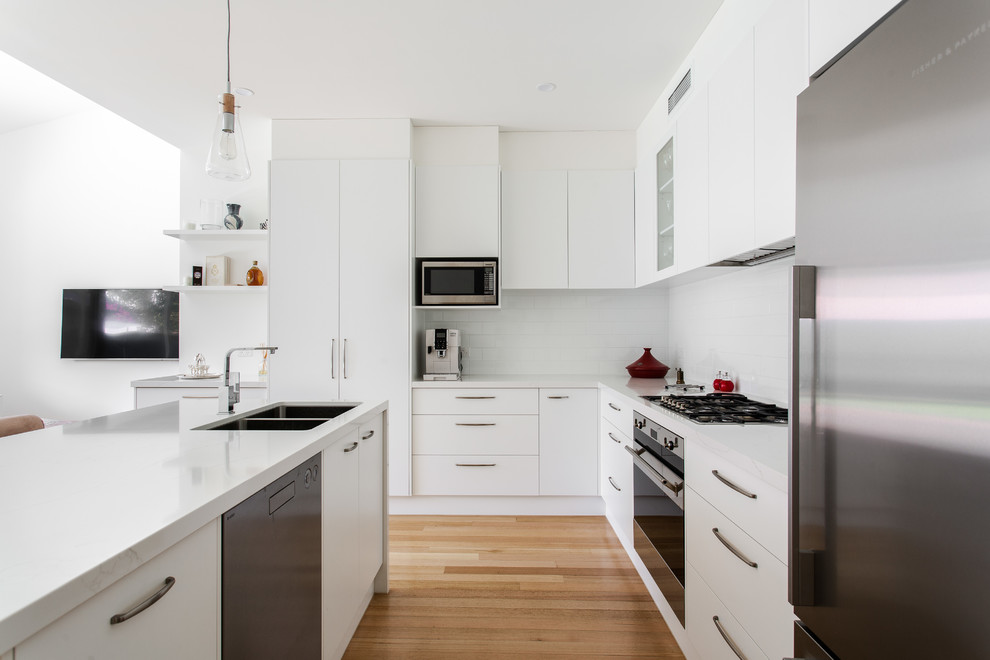 Inspiration for a contemporary u-shaped kitchen in Adelaide with a double-bowl sink, flat-panel cabinets, white cabinets, white splashback, subway tile splashback, stainless steel appliances, medium hardwood floors, a peninsula, brown floor and white benchtop.