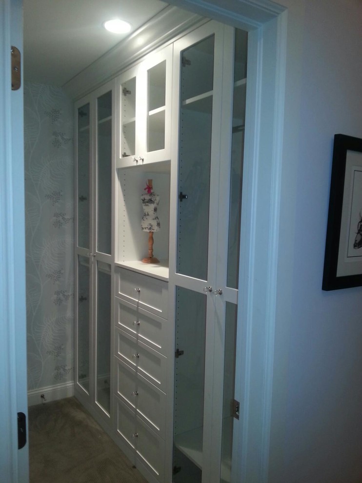 Mid-sized traditional gender-neutral walk-in wardrobe in New York with glass-front cabinets, white cabinets, carpet and beige floor.