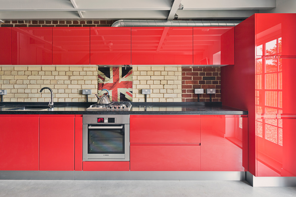This is an example of a small industrial single-wall open plan kitchen in Cambridgeshire with a drop-in sink, flat-panel cabinets, red cabinets, brick splashback, stainless steel appliances, concrete floors and grey floor.