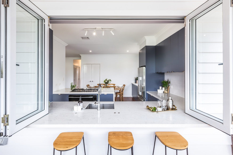 This is an example of a small transitional l-shaped eat-in kitchen in Brisbane with an undermount sink, flat-panel cabinets, blue cabinets, quartz benchtops, white splashback, subway tile splashback, black appliances, medium hardwood floors, with island, brown floor and white benchtop.
