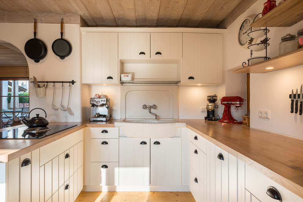 Small country eat-in kitchen in Munich with a farmhouse sink, louvered cabinets, white cabinets, wood benchtops, white appliances, medium hardwood floors, a peninsula, brown floor and brown benchtop.