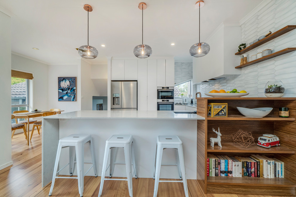 Photo of a mid-sized contemporary u-shaped open plan kitchen in Canberra - Queanbeyan with an undermount sink, white cabinets, quartz benchtops, grey splashback, stainless steel appliances, light hardwood floors, white benchtop, flat-panel cabinets, no island and brown floor.
