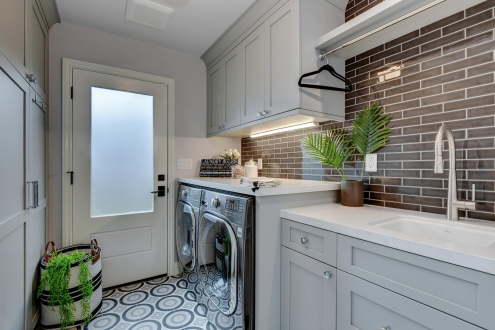 Design ideas for a mid-sized contemporary galley dedicated laundry room in San Francisco with an undermount sink, raised-panel cabinets, grey cabinets, quartz benchtops, grey walls, ceramic floors, a side-by-side washer and dryer, multi-coloured floor, grey benchtop, black splashback and glass sheet splashback.