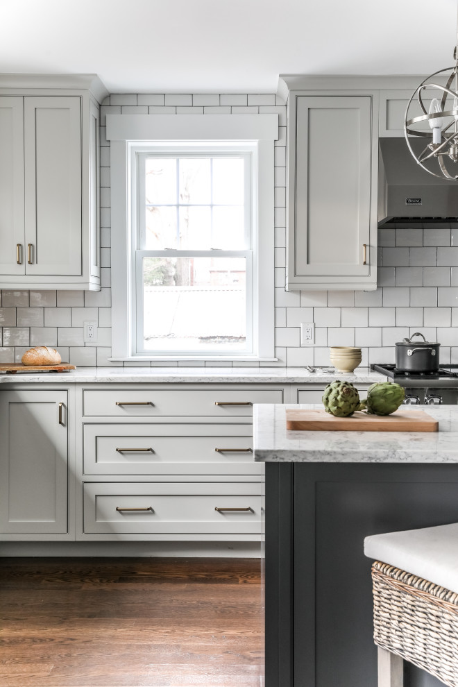 Design ideas for a transitional l-shaped eat-in kitchen in Boston with a farmhouse sink, beaded inset cabinets, white cabinets, quartz benchtops, white splashback, subway tile splashback, stainless steel appliances, medium hardwood floors, with island, brown floor and grey benchtop.