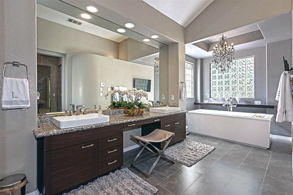 This is an example of a mid-sized contemporary master bathroom in Las Vegas with recessed-panel cabinets, dark wood cabinets, a freestanding tub, a double shower, a one-piece toilet, glass sheet wall, grey walls, ceramic floors, a drop-in sink, granite benchtops, grey floor, a hinged shower door and multi-coloured benchtops.