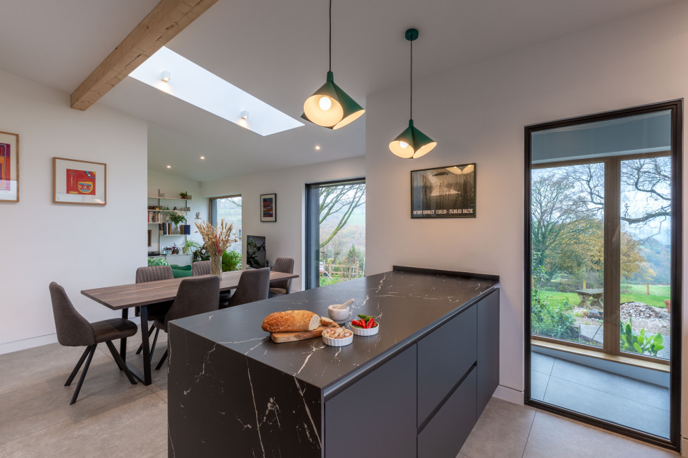 This is an example of a medium sized industrial l-shaped open plan kitchen in Cheshire with a double-bowl sink, flat-panel cabinets, grey cabinets, composite countertops, grey splashback, mirror splashback, black appliances, porcelain flooring, an island, grey floors, black worktops, exposed beams and feature lighting.
