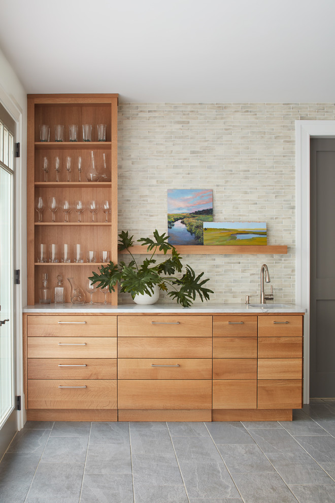 This is an example of a beach style single-wall wet bar in Boston with an undermount sink, flat-panel cabinets, medium wood cabinets, beige splashback, grey floor and white benchtop.