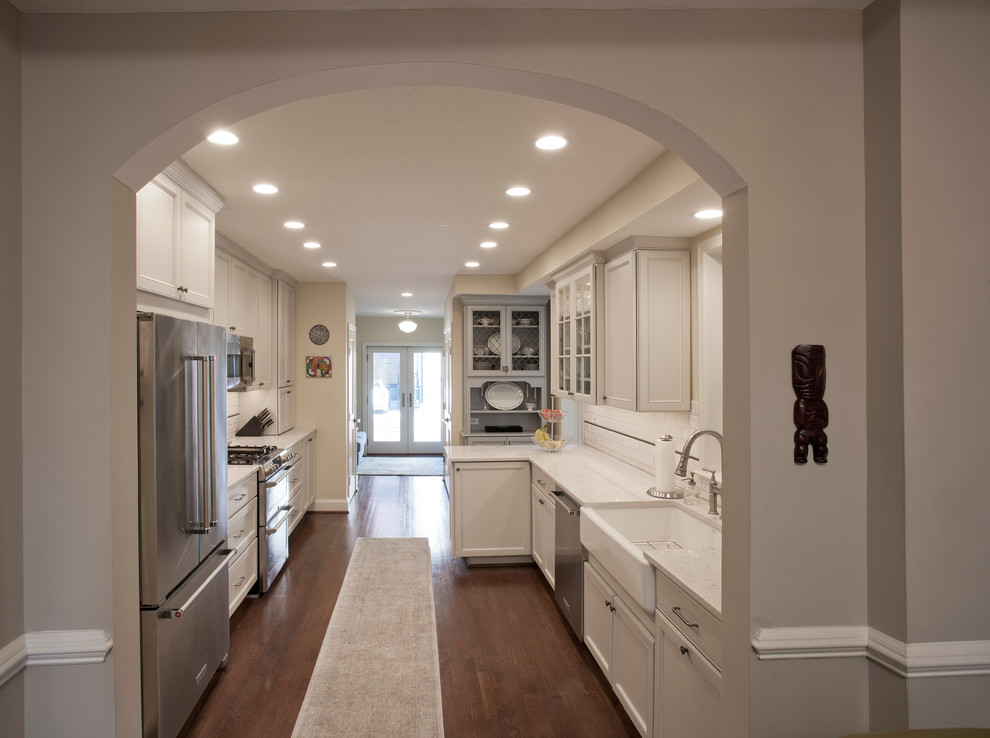 Photo of a traditional galley separate kitchen in DC Metro with a farmhouse sink, recessed-panel cabinets, quartz benchtops, white splashback, subway tile splashback, stainless steel appliances, dark hardwood floors, a peninsula and grey cabinets.