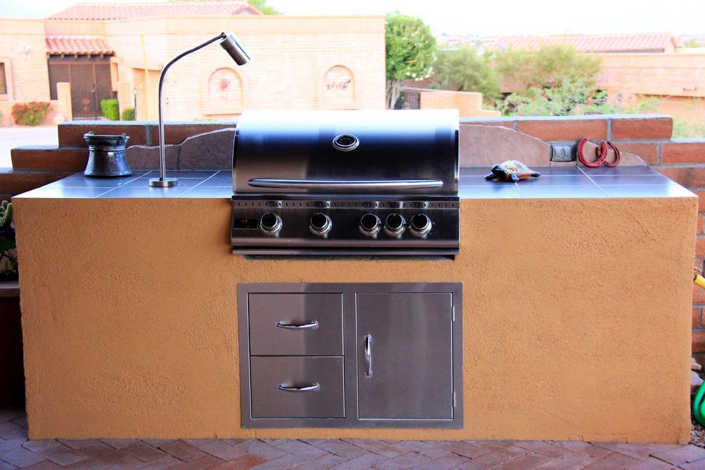 This is an example of a mid-sized backyard patio in Phoenix with an outdoor kitchen, brick pavers and a roof extension.