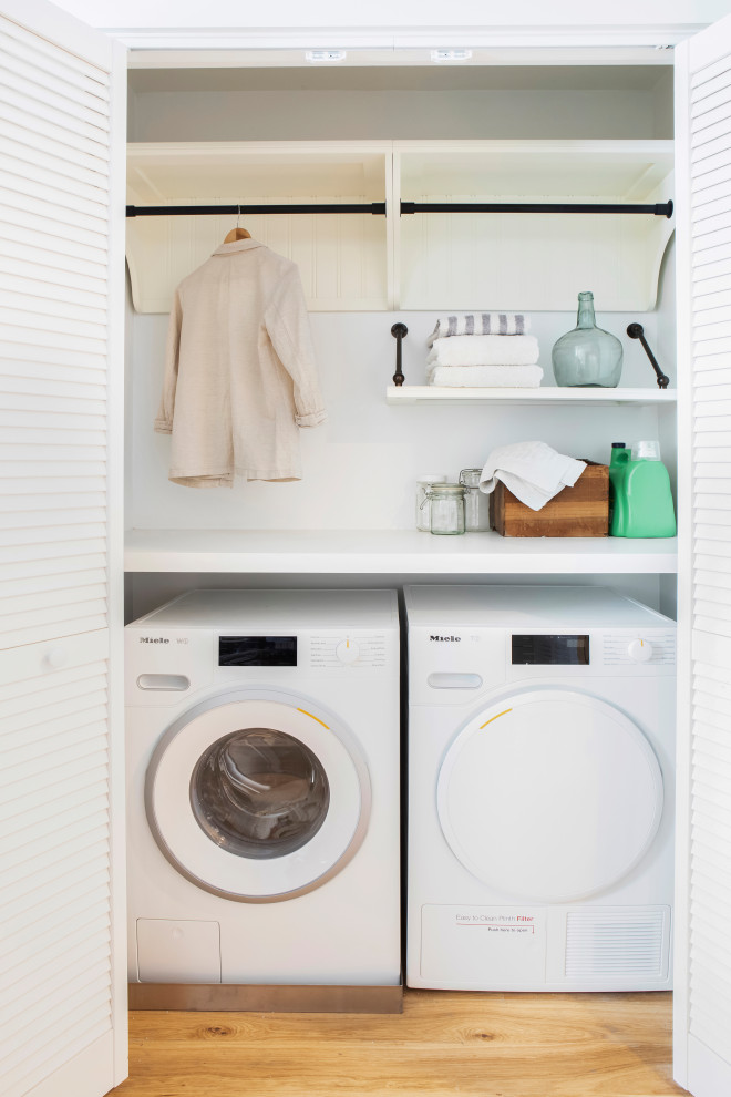 This is an example of a small contemporary single-wall laundry cupboard in New York with open cabinets, white cabinets, solid surface benchtops, white walls, medium hardwood floors, a side-by-side washer and dryer, brown floor and white benchtop.