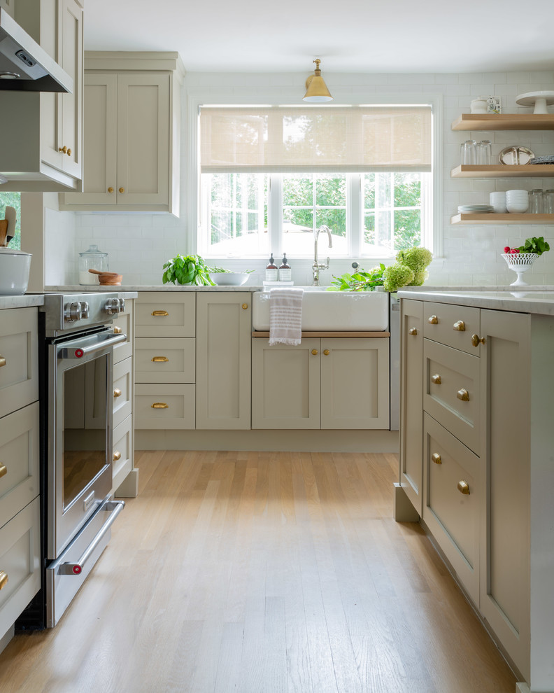 Mid-sized transitional l-shaped kitchen in Boston with a farmhouse sink, shaker cabinets, beige cabinets, marble benchtops, white splashback, subway tile splashback, stainless steel appliances, light hardwood floors, with island, brown floor and white benchtop.