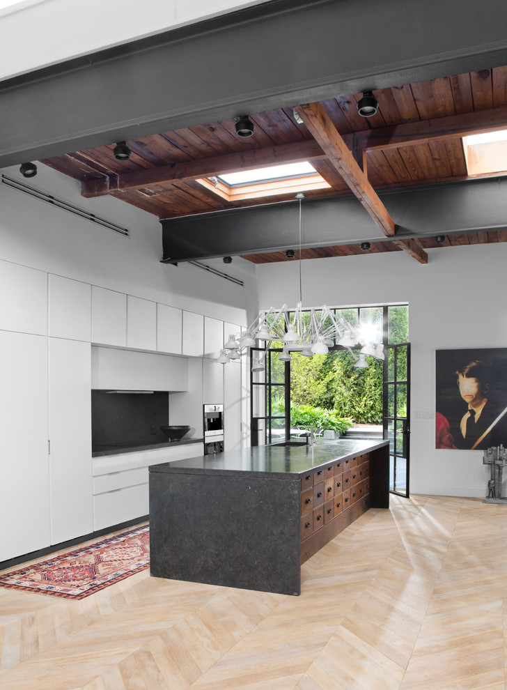 Photo of a large industrial single-wall open plan kitchen in Other with an undermount sink, flat-panel cabinets, white cabinets, limestone benchtops, black splashback, panelled appliances, light hardwood floors, with island and brown floor.