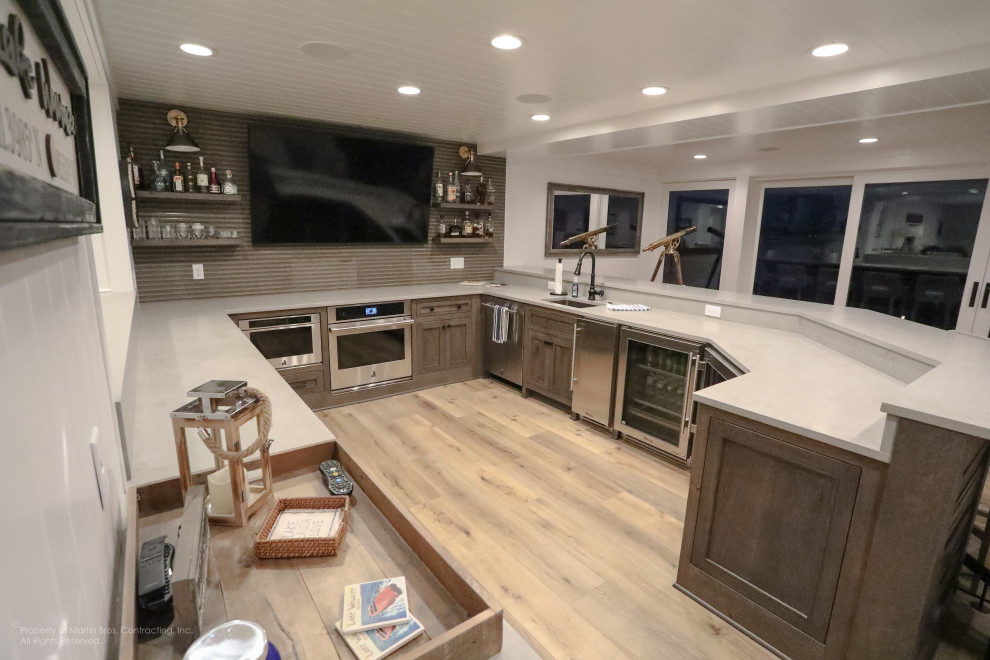 Design ideas for an expansive beach style u-shaped seated home bar in Other with an undermount sink, flat-panel cabinets, grey cabinets, quartz benchtops, grey splashback, matchstick tile splashback, medium hardwood floors and grey benchtop.