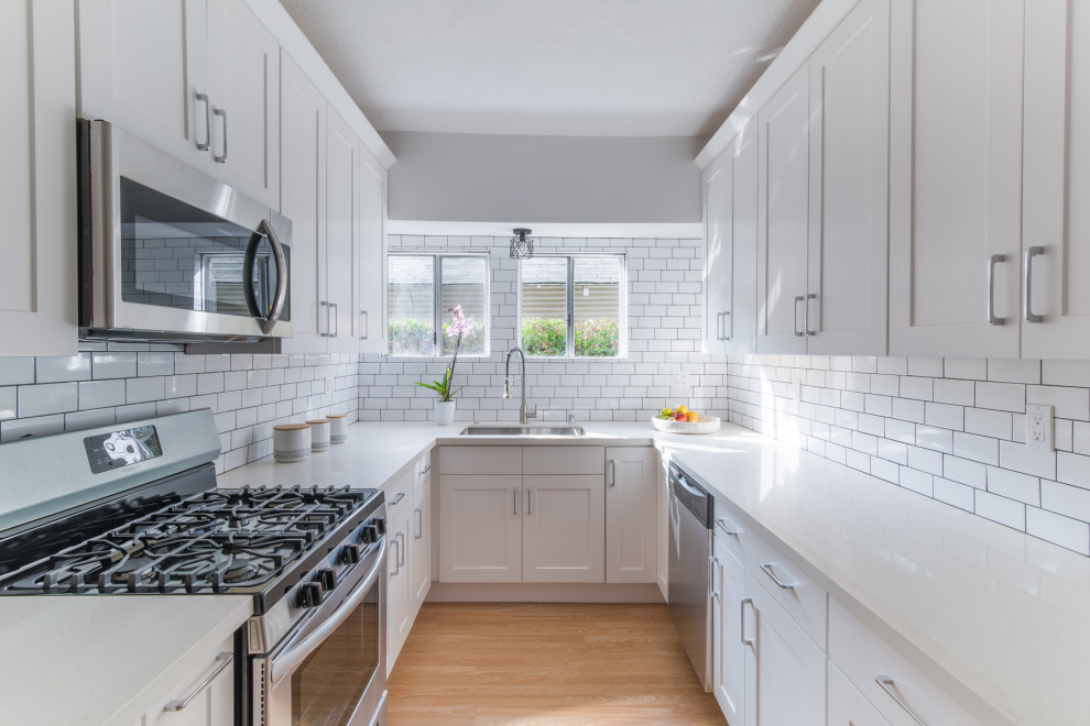 Photo of a small contemporary galley kitchen in Los Angeles with an undermount sink, shaker cabinets, white cabinets, solid surface benchtops, white splashback, subway tile splashback, stainless steel appliances, light hardwood floors, no island, brown floor and beige benchtop.
