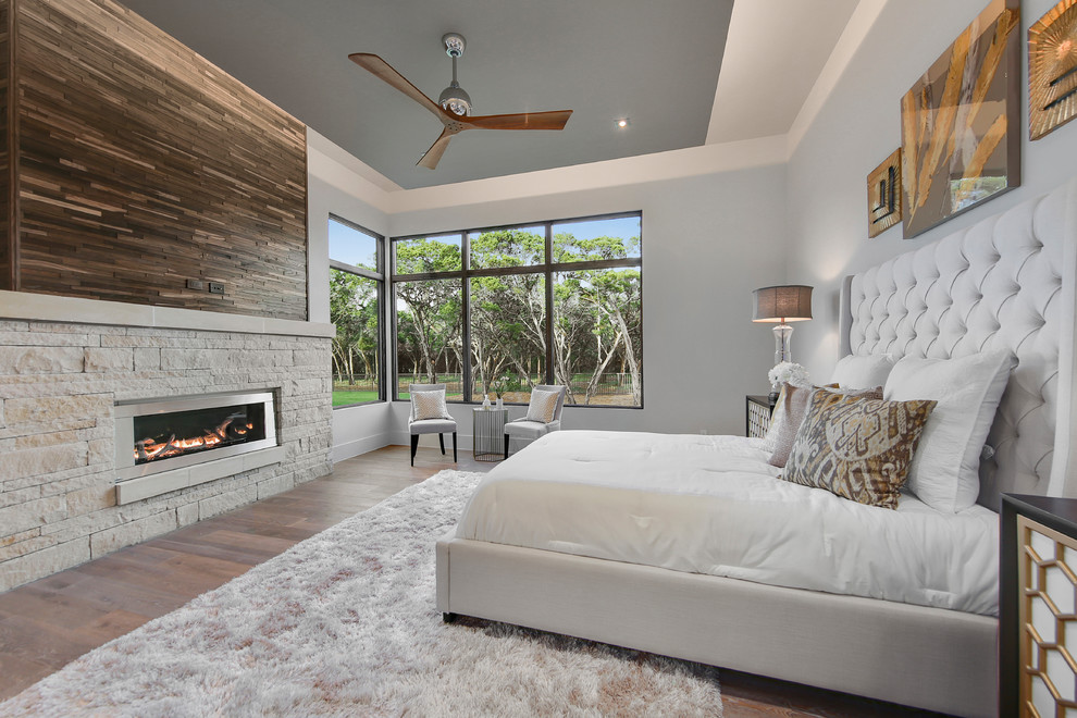 Large transitional master bedroom in Austin with grey walls, light hardwood floors, a ribbon fireplace, a stone fireplace surround and brown floor.