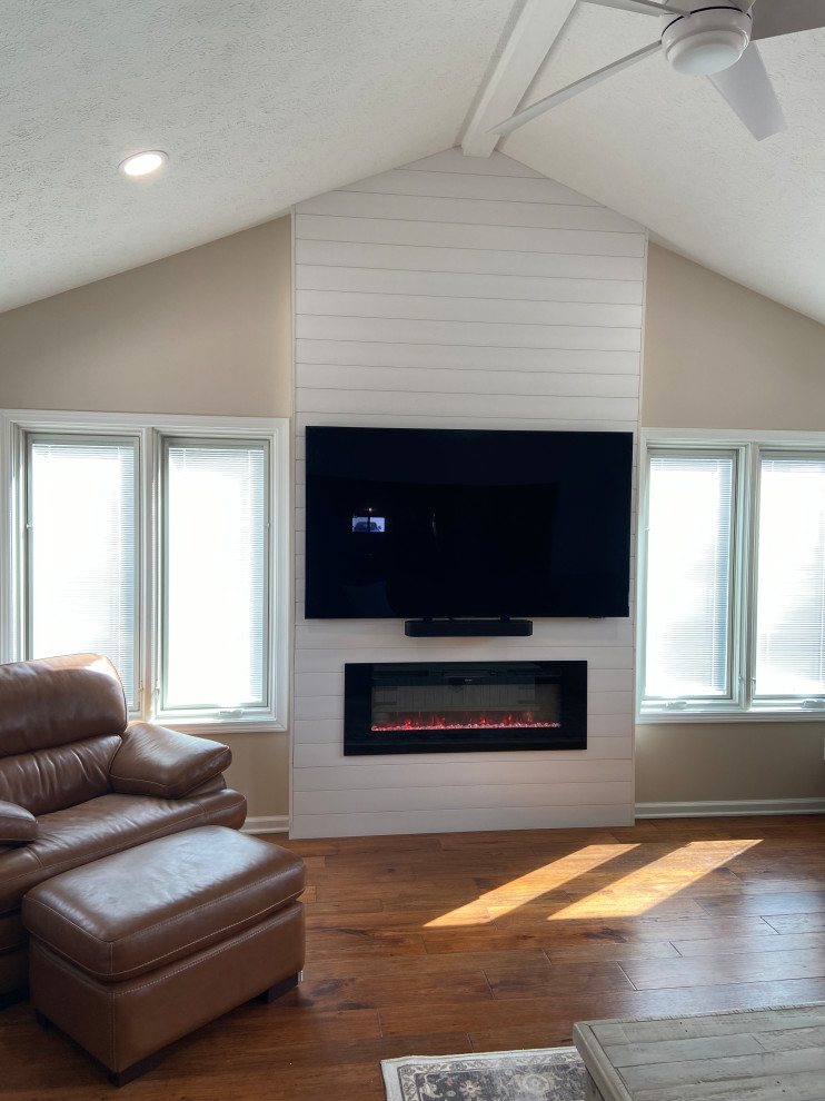 Inspiration for a large rural open plan games room in Indianapolis with beige walls, medium hardwood flooring, a hanging fireplace, a timber clad chimney breast, a wall mounted tv, beige floors and a vaulted ceiling.