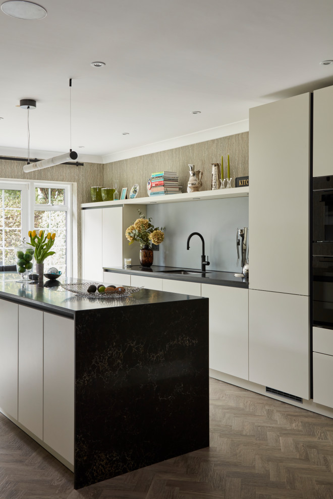 Inspiration for a medium sized contemporary cream and black single-wall enclosed kitchen in London with a submerged sink, flat-panel cabinets, white cabinets, engineered stone countertops, grey splashback, glass sheet splashback, black appliances, medium hardwood flooring, an island, brown floors, black worktops and feature lighting.
