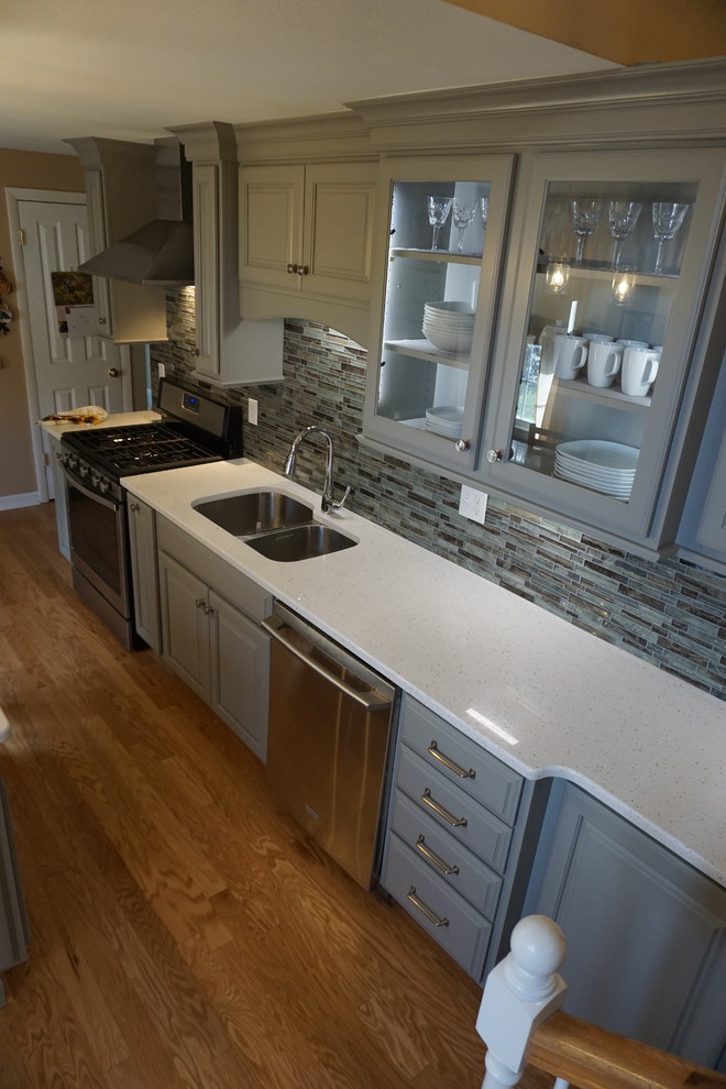 Example of a small classic l-shaped medium tone wood floor eat-in kitchen design in Providence with a triple-bowl sink, raised-panel cabinets, gray cabinets, quartz countertops, gray backsplash, mosaic tile backsplash, stainless steel appliances, an island and white countertops